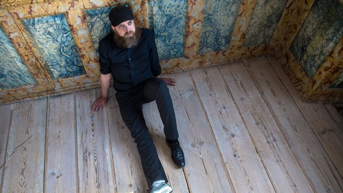 Mand med lang barbe sidder på gulv i lille rum med blå og gule mønstre på vægge