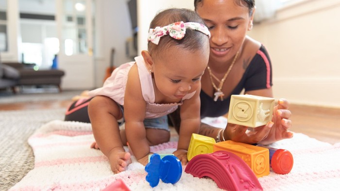 Ein Baby spielt mit bunten Spielzeugen auf einer Decke, während eine Frau daneben kniet.