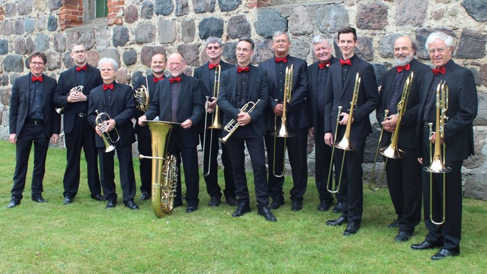 Eine Gruppe von Musikern posiert vor einer Steinmauer, alle halten Blasinstrumente und tragen schwarze Anzüge mit roten Einstecktüchern.