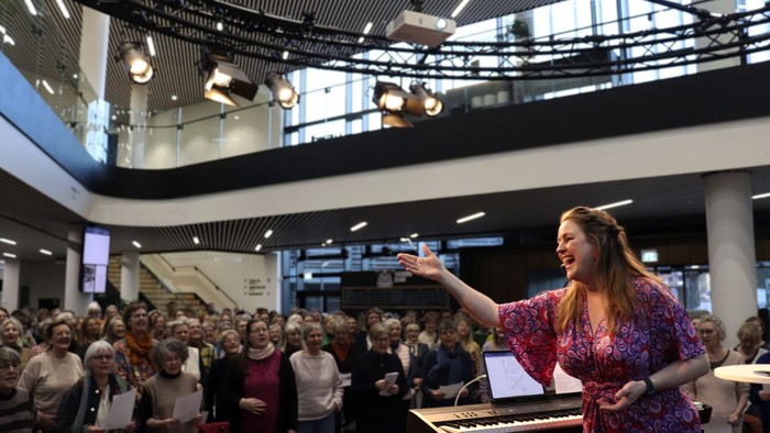 En kvinde på scenen med mikrofon, tale til et publikum i et moderne lokale.