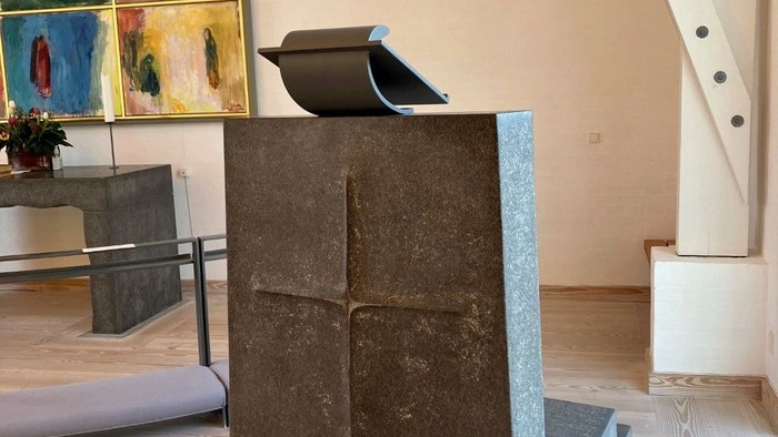 En stor sten med et udhugget kors står i et rum med billeder og trappe.