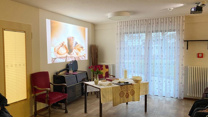 Wohnzimmer mit Holzboden, rotem Sessel, Leinwand für Beamer und Tisch mit Gegenständen.