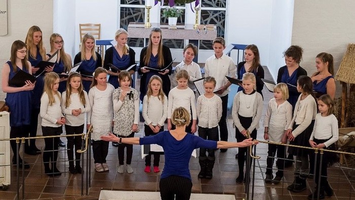 En gruppe børn og voksne står i en hal og synger i kor.