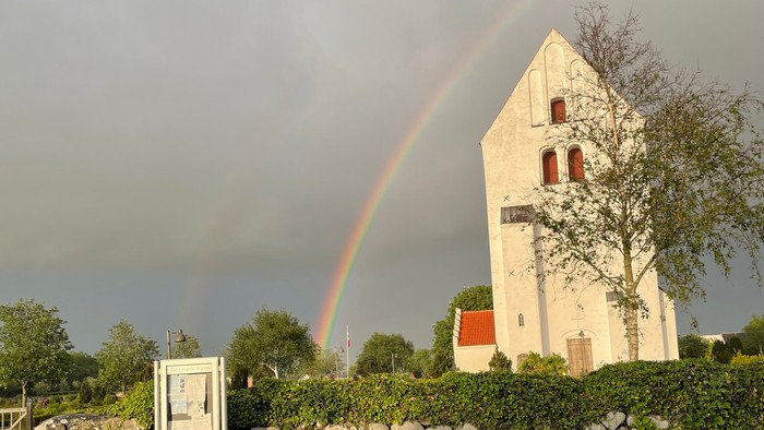 Regnbuen spænder over kirke med blå bil og stenmur foran