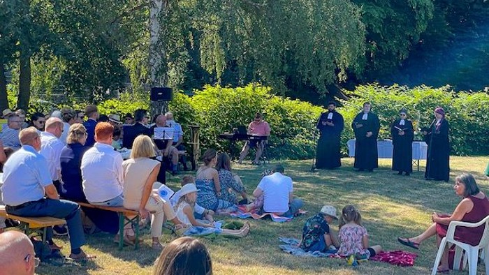 Eine Gruppe von Menschen sitzt im Freien auf Bänken und Decken, umgeben von Bäumen, und hört einer Gruppe von Personen zu, die auf einer Bühne stehen.