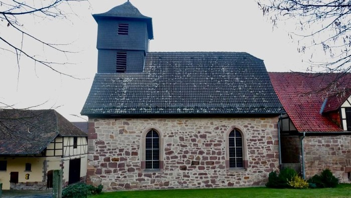 Kleine Steinkirche mit Glockenturm und rot gedecktem Gebäude.