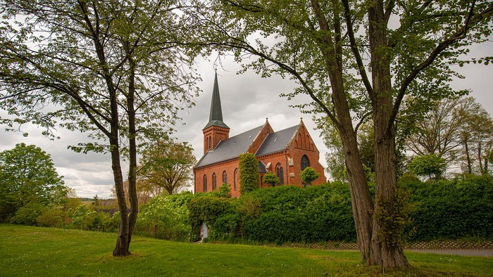 Ein rotes Ziegelgebäude mit spitzem Turm steht in einem grünen Park.