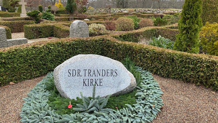En mindesten med indskrivet "SDR.TRANDERS KIRKE" i en parkagtig omgivelse med buske og træer.