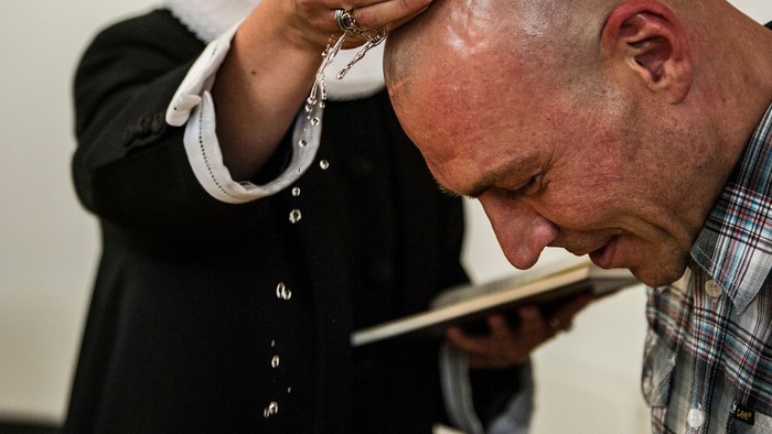 En bald mand læser fra en bog mens en anden person holder en rosary ved hans hoved.