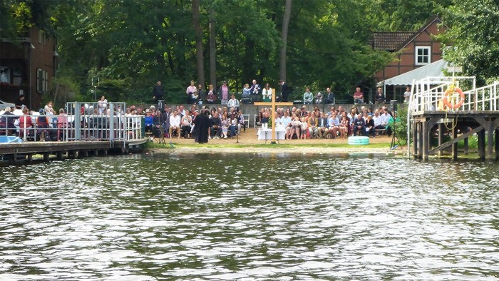 Eine Gruppe von Menschen versammelt sich am Ufer eines Flusses, einige stehen auf einem Steg.