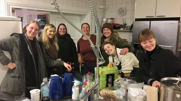 En gruppe af venner køkkenet, hvor de smiler og står ved spisebordet med mad og drikkevarer.