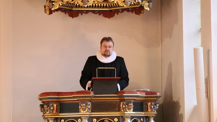 Mand i sort kappe og halsbånd står ved prædikestol i kirke