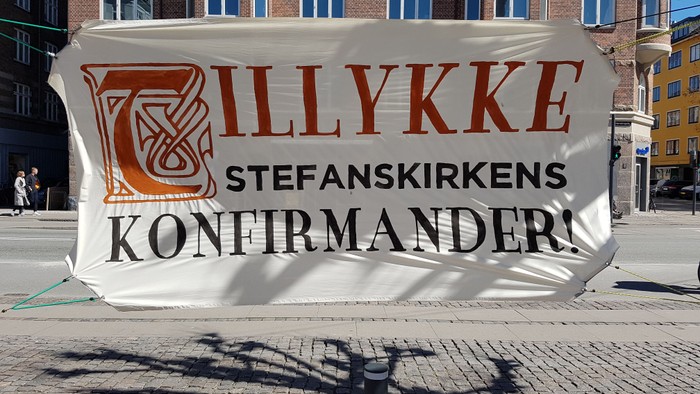 Stor banner med teksten "ILLYKKE STEFANSKIRKENS KONFIRMANDER" på en bygning