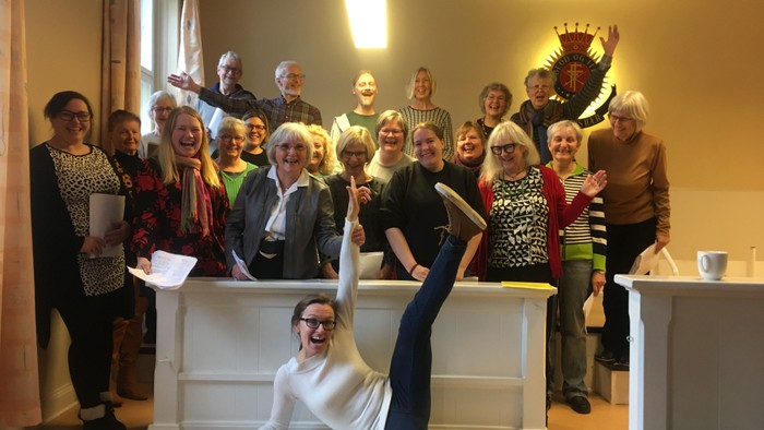 En gruppe af ældre personer samles i et rum, hvor en kvinde laver en akrobatisk pose på en bord.