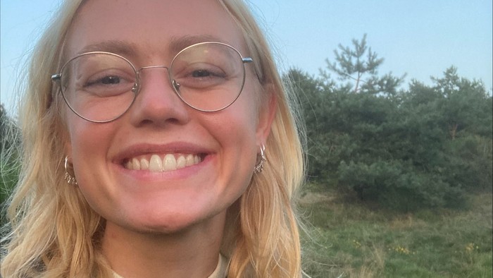 En ung kvinde med blonder og briller smiler til kameraet udendørs.