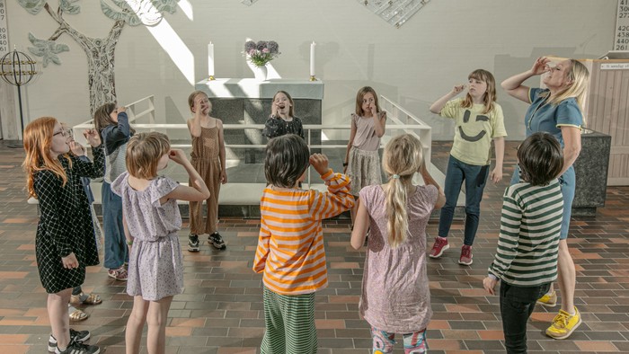 En gruppe børn og voksne samles i et rum med muralske vægge og træfliser.