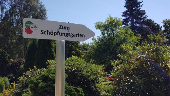 Weißes Schild mit grüner Beschriftung zeigt Richtung "Zum Schöpfungsgarten" in einem Garten.