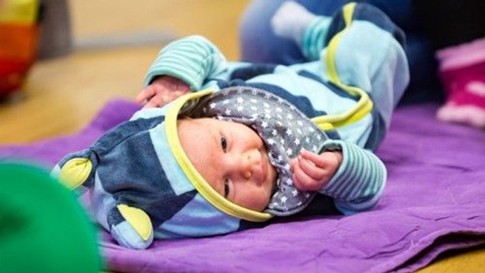 Et spæd barn ligger på en violet duk og smiler, mens det holder en gule ring.