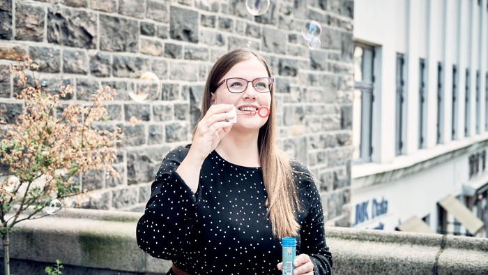En ung pige med briller holder en blå flaske og smiler ved en mur.