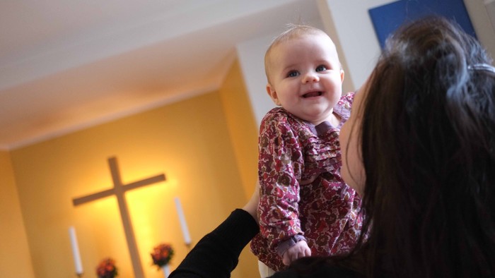 Et barn i rødt og hvitt mønsteret skjorte holdes oppe af en voksne, mens de står i en kirke.