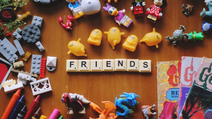 Scrabble tiles spell "FRIENDS" surrounded by various toys and crayons on a wooden surface.