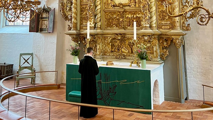 En præst i svart kappe står foran en prægtig altertavle i en kirke.