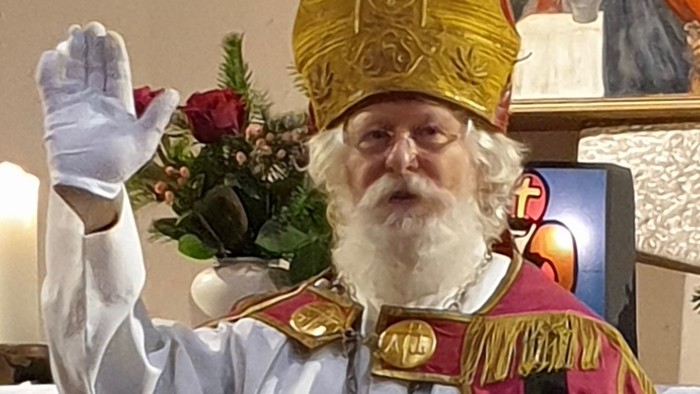 Ein Mann in kirchlicher Kleidung mit goldenem Hut und weißem Bart hält eine Blume hoch