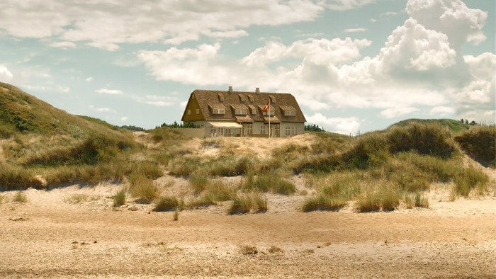 En stor, træklædt husstand på en sandstrand med græs og klodebjerge.