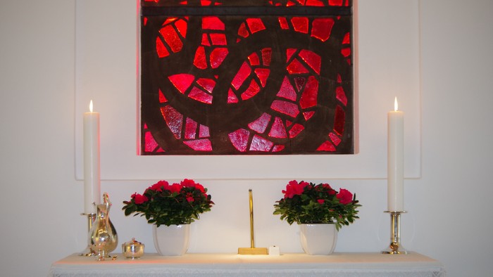 En altertavle med to lyse lysestake, to blomsterstærke og et rødt glassmaleri.