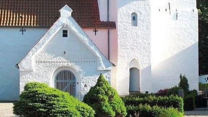 En smuk og hvid kirke med et trægt tæt på og en have med buske og blomster.