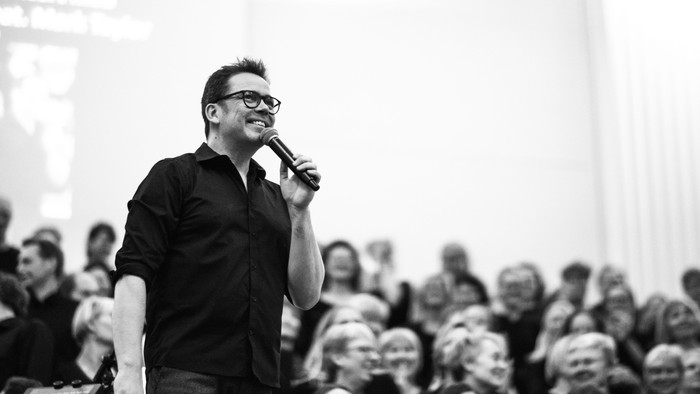 En mand med briller holder mikrofon og taler til et publikum.