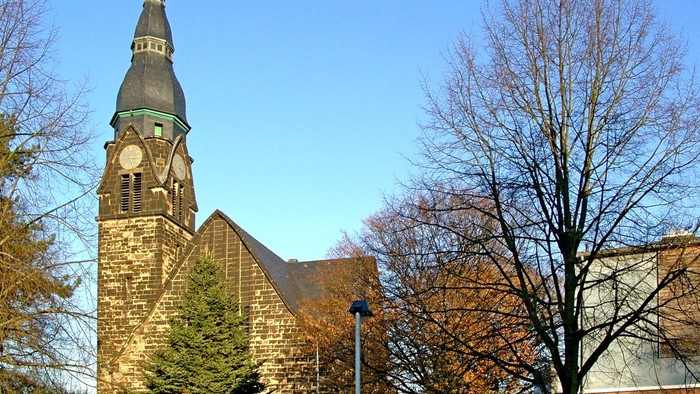 Kirchturm mit Uhr an der Christuskirche aus dunklem Stein, umgeben von Herbstbäumen ohne Blätter und einem klaren blauen Himmel.