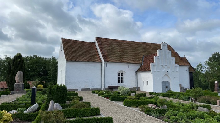 Et hvidt kirkebygning med røde tag og en gravplads med høje buske og graver.