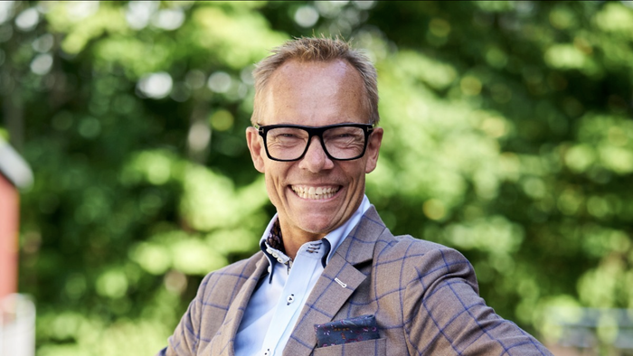 En smilende mand med briller og flau hår, der bærer en fløjteblå og grå jakke.