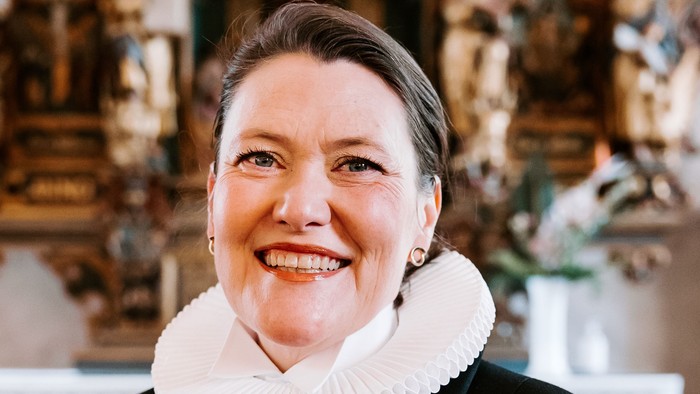 En kvinde i sort jakke med hvid krage står foran en prægtig altertavle.