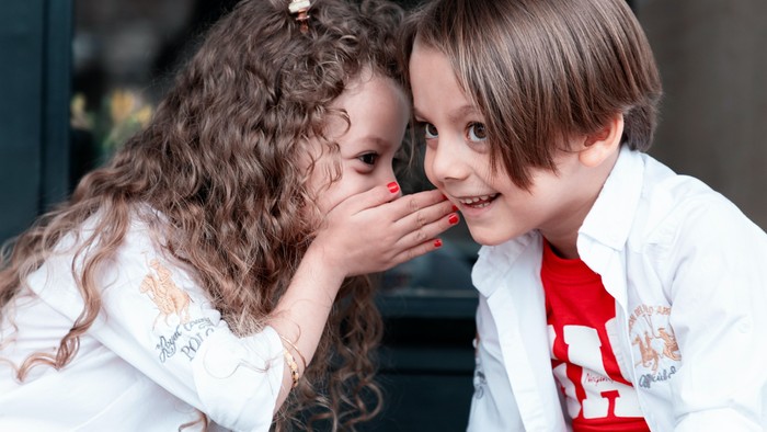 Ein Mädchen mit lockigem Haar flüstert einem Jungen mit kurzem Haar etwas ins Ohr, beide lächeln.