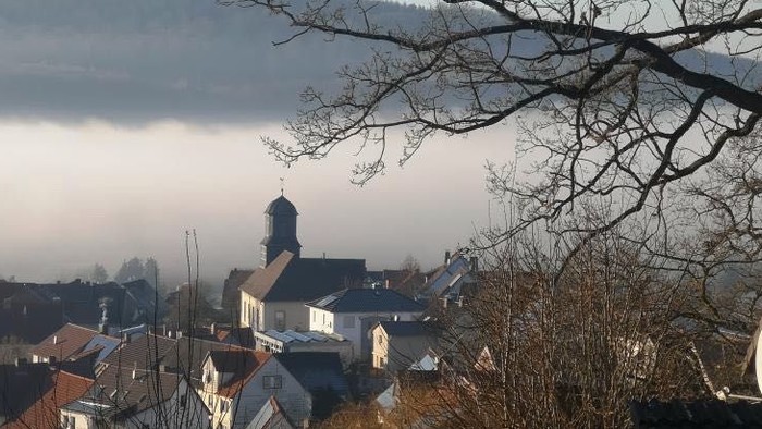 Neblige Stadt mit Kirchturm, umgeben von Hügeln und kahlen Bäumen.