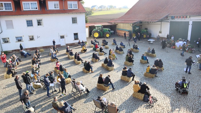 Menschen sitzen auf Heuballen auf einem Hof mit Gebäuden und Traktoren.