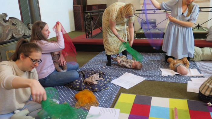 En gruppe af voksne og børn sidder på gulvmatte og spiller med farver og papir.