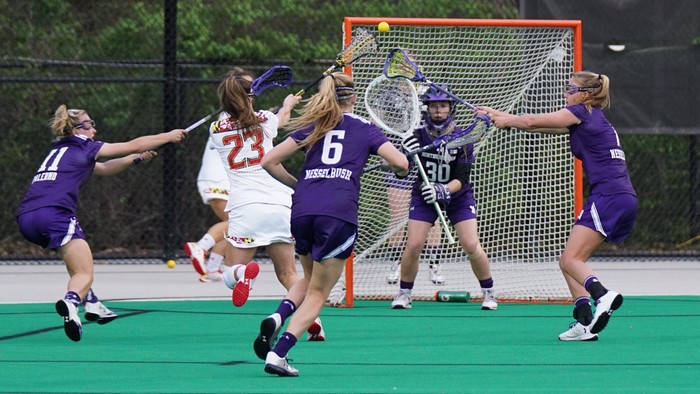 Frauen-Lacrossespielerinnen in Aktion vor dem Tor: Eine Spielerin in Weiß wirft den Ball, Verteidigerinnen in Lila versuchen zu blocken.