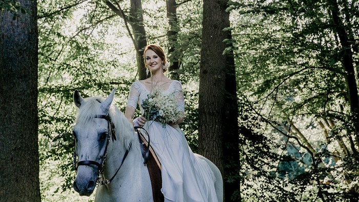 Eine Frau in Hochzeitskleid reitet auf einem weißen Pferd durch einen Wald.