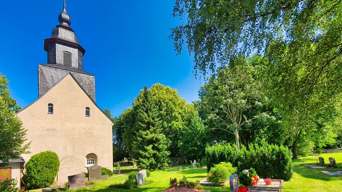 Ein weißer Kirchturm mit schwarzer Spitze steht in einem grünen Park mit Bäumen und Bänken.