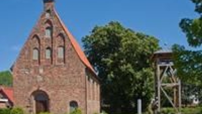 Ein rotes Backsteingebäude mit spitzem Dach und einem Schornstein, umgeben von Bäumen und einem Zaun.