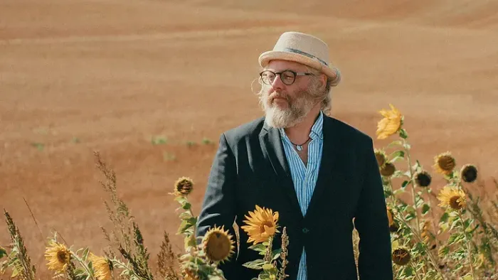 En bjergmand med hat og briller står i en solrosenmark med rød jord i baggrunden