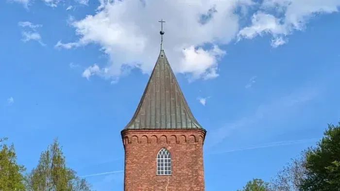 Kirchturm aus rotem Backstein vor blauem Himmel und Bäumen