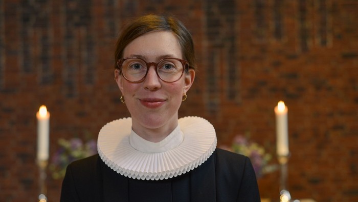 Kvinde med briller og hvid krage står forud for mur med lysede lygter