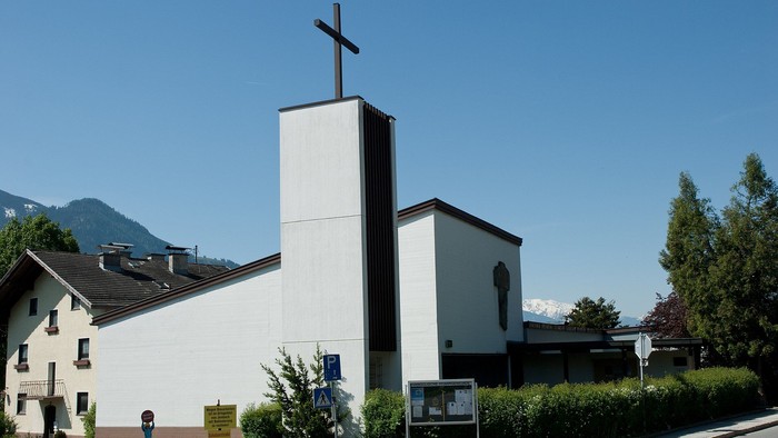 Weiße Kirche mit hohem Turm und Kreuz, umgeben von Grün, im Hintergrund Berge