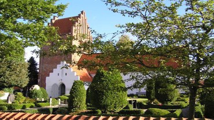 Et rødt teglsteinskirke med høje tårne omgivet af grønne træer og buske