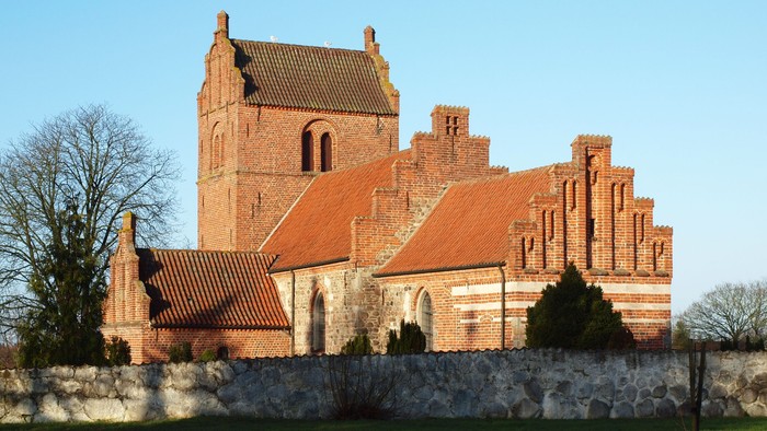 Gamle kirke med røde tagtegnere og mursten, omgivet af træer og en mur.