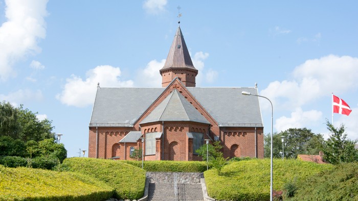 En rød kirke med spiret og en dansk flagvåbens flag på en klar dag